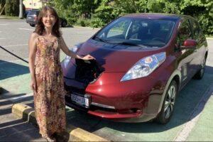 A woman stands next to a red electric car as it’s connected by a charging cable to a street charger.