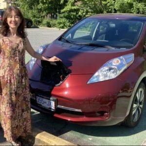 A woman stands next to a red electric car as it’s connected by a charging cable to a street charger.