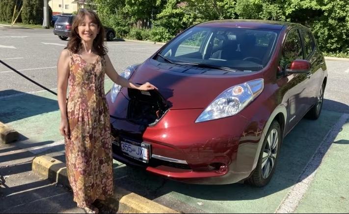 A woman stands next to a red electric car as it’s connected by a charging cable to a street charger.